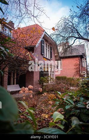 A two-story brick house with large windows and wooden shutters. A bench overlooking an autumn garden and trees with yellowing leaves. Stock Photo