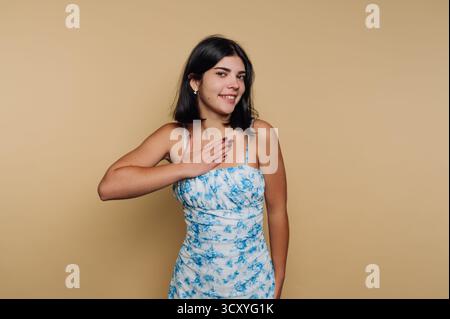 A young woman stands smiling, her hand touching her chest in a friendly gesture. She wears a blue floral dress and poses confidently against a warm, s Stock Photo