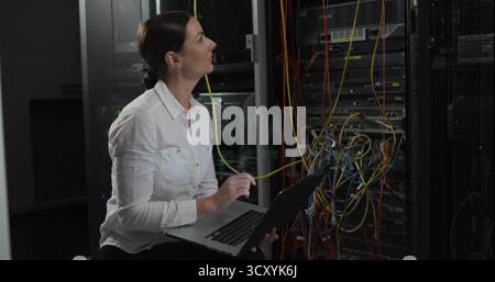 Kneeling IT technician tracing yellow Ethernet cable beside server rack in data center with laptop Stock Photo