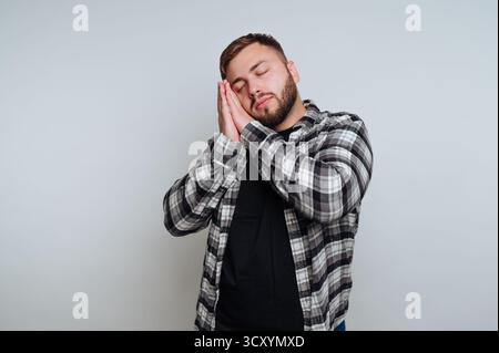 A man stands with his eyes closed, cradling his face as if resting or feeling sleepy. He wears a plaid shirt over a black t-shirt, set against a simpl Stock Photo