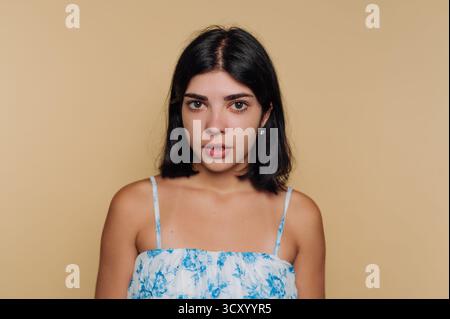 A woman stands in front of a plain beige background, displaying a neutral expression. She wears a light floral dress and has shoulder-length black hai Stock Photo