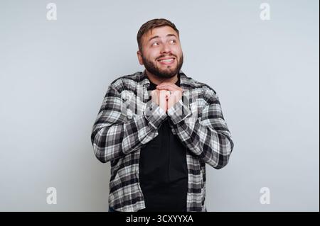 A man wearing a checkered shirt with a joyful expression stands in front of a plain backdrop. His hands are clasped together as he looks hopeful and c Stock Photo