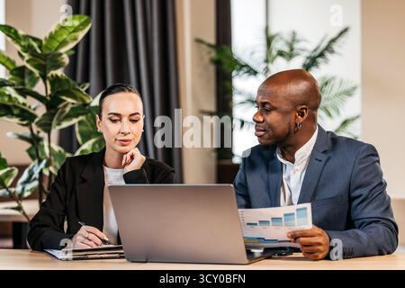 Business professionals collaborating in a modern office, analyzing financial data on a laptop, with plants and contemporary decor enhancing the worksp Stock Photo