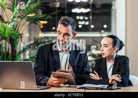 Two business professionals are discussing strategies in a modern office, using a laptop and tablet, with plants and stylish decor enhancing the worksp Stock Photo