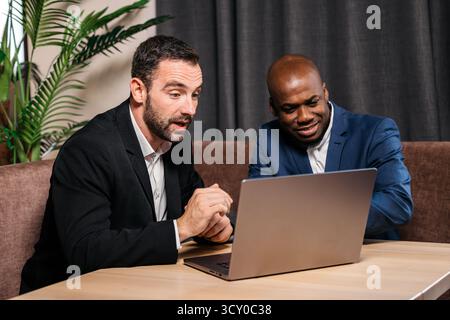 Two men are collaborating over a laptop in a contemporary workspace, surrounded by plants, highlighting teamwork, focus, and modern business dynamics Stock Photo