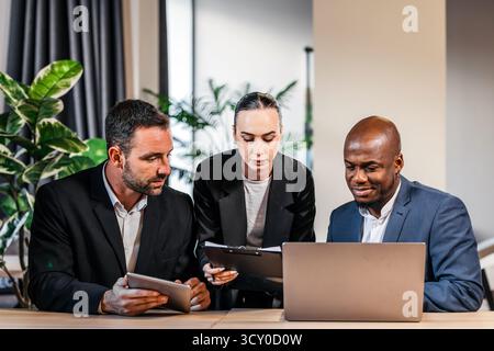 Diverse team members engaged in collaboration at a contemporary office, reviewing project details on digital devices and a laptop, highlighting teamwo Stock Photo