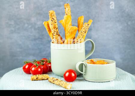 Bread sticks with seeds in the ceramic mug, tomatoes and cheese sauce. Homemade bread sticks in the cup on gray background and green textile. Stock Photo
