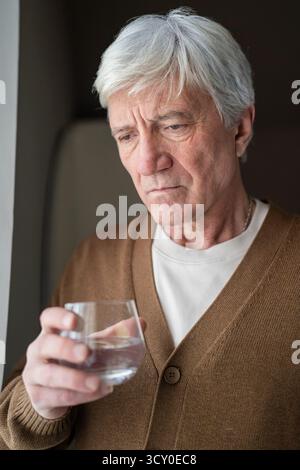 Unhappy elderly man drink water from glass in solitary, aging depression. Melancholic male grief Stock Photo
