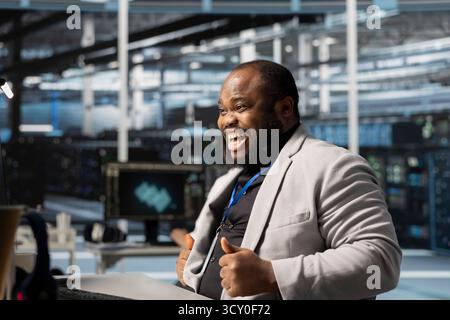 Overjoyed programmer in server hub satisfied by improved system efficiency metrics. Ecstatic African american man working in data center pleased by enhanced network infrastructure performance Stock Photo