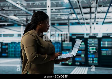 Woman in data center conducting tests on infrastructure using software to fix bugs. African american server hub worker using laptop to refine algorithms for faster processing, updating core services Stock Photo