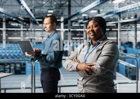 Portrait of smiling server room engineer next to coworker troubleshooting errors using software on laptop. Cheerful african american woman in data center and colleague fixing bugs using notebook Stock Photo