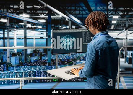 Black female IT specialist reviews AI performance data and LLM visualizations on computer workstation. Engineer overseeing neural network for server hub infrastructure operations. Stock Photo