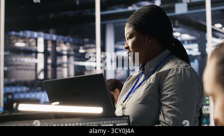 Data center manager supervising engineer developing software to analyze neural networks at computer workstation. Team leader and employee observing system performance in AI server room, camera A Stock Photo