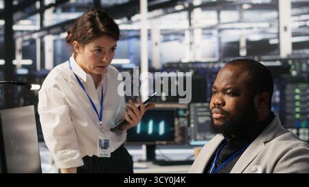 Lead data center admin overseeing IT expert integrating AI in equipment rigs. African american employee under server farm team leader supervision using deep learning for operational tasks, camera A Stock Photo