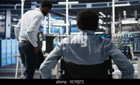 Data center programmer in wheelchair entering office uses AI node tree program to minimize system downtime. Server room IT expert in disability uses machine learning visual programming tool, camera A Stock Photo