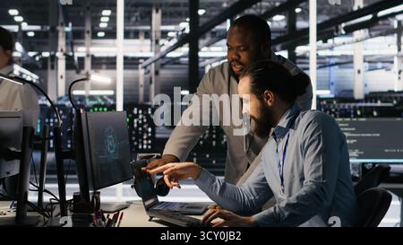 Data center engineers use laptop, collaborating on doing AI software updates. Server room teamworking colleagues doing brainstorming, optimizing infrastructure using artificial intelligence, camera A Stock Photo