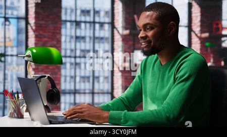 African american male teleworker completing spreadsheets on laptop, entering numerical data into cells and calculating totals. Guy using spreadsheet software functions. Camera A. Stock Photo