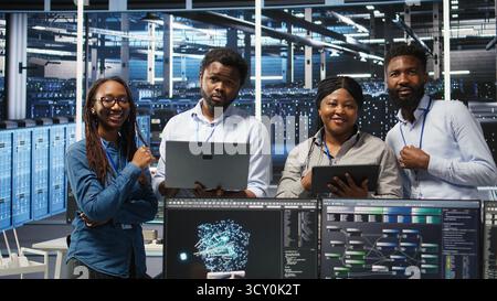 Portrait of smiling data center specialists collaborating in server facility, using node tree software to analyze datasets. Happy team using visual programming to improve gear performance, camera A Stock Photo