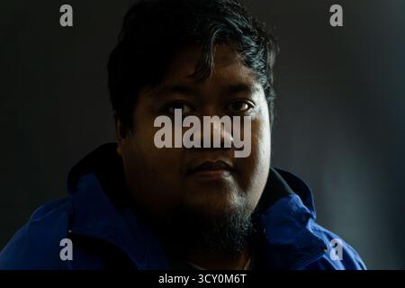 Bearded Indonesian man in his 30s wearing a shiny blue nylon jacket against black background, casual portrait with confident expression and simple lig Stock Photo