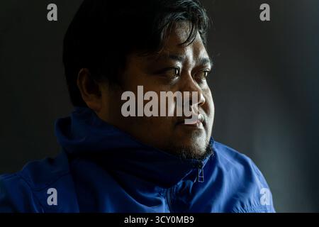Bearded Indonesian man in his 30s wearing a shiny blue nylon jacket against black background, casual portrait with confident expression and simple lig Stock Photo