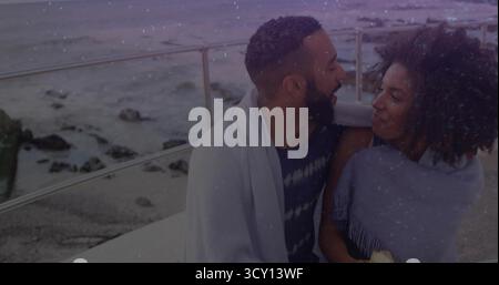 Smiling couple wrapped in blankets sitting on seaside ledge with metal railing, holding ceramic mug Stock Photo