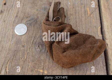 A rustic brown suede drawstring pouch on a weathered wooden surface with a silver coin for scale Stock Photo
