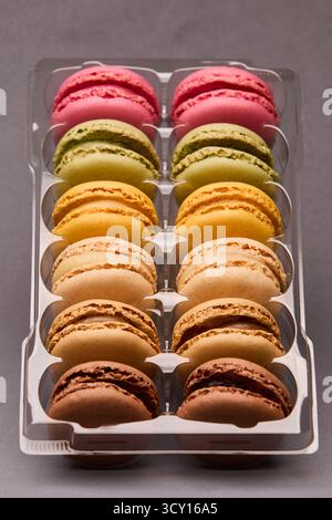Vertical shot of bright assorted macarons neatly arranged in plastic box on gray background. Modern patisserie presentation with color harmony. Stock Photo