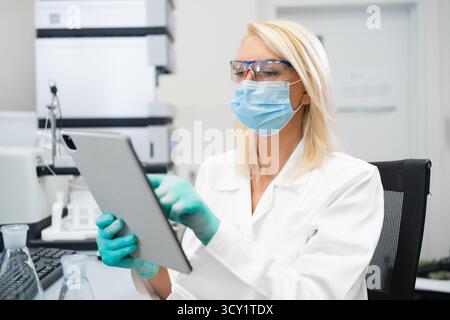 Female scientist in lab coat and mask using tablet to analyze data in laboratory. Stock Photo