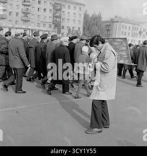 An archival photo from November 7, 1974, captures a candid moment during an October Revolution parade in Sloviansk. A young amateur photographer, fashionably dressed for the era, stands on the sidelines, checking his vintage film camera while holding balloons. In the background, a column of demonstrators marches past. This image provides a unique perspective on the event, focusing on an individual documenting the celebration. It's a nostalgic snapshot of personal hobbies and public life in a peaceful Donbas before the war Stock Photo