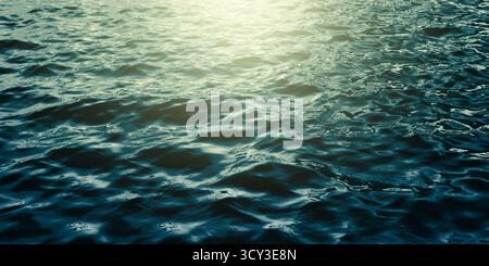 Dark, rippling water sea dramatic. Surface is covered in small waves. Bright, golden-yellow light shines down. Water color transitions from deep teal Stock Photo