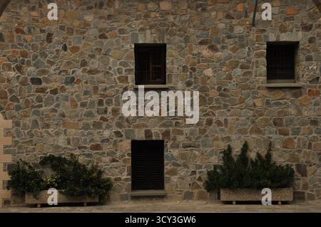 A textured stone wall showcases three rustic windows and two planters with lush green plants. The scene is calm and invites a sense of tranquility and Stock Photo