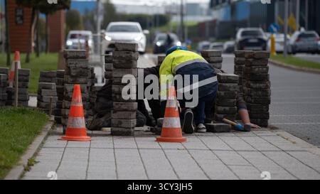 Road maintenance workers repairing a sidewalk in an industrial area surrounded by traffic cones and brick stacks. The image captures everyday urban in Stock Photo