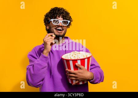 Cheerful man in a stylish hoodie smiling while holding a large popcorn bucket on a vibrant yellow background Stock Photo