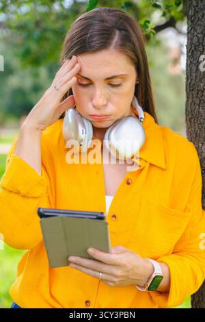 Young caucasian woman frustrated while using digital tablet outdoors in park Stock Photo