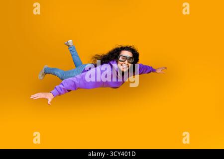 Funky man in casual hoodie smiling and posing mid-air on vibrant yellow backdrop, embracing joyful energy and style. Stock Photo