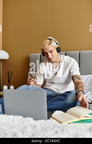 Handsome young man with headphones sits on bed, focused on laptop while taking notes. Stock Photo