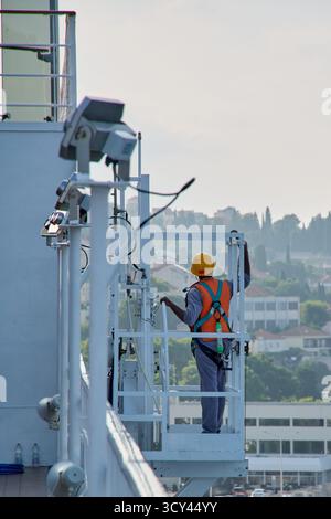 Dubrovnik. Croatia - October 17, 2025: An industrial worker seen from behind, wearing an orange vest, helmet, and safety harness, performs maintenance Stock Photo