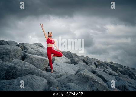 Woman wearing red sportswear doing a yoga tree pose on rugged rocks Stock Photo