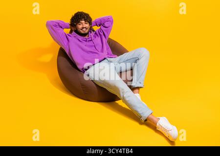 Young man relaxing on a beanbag chair against a yellow background wearing casual purple hoodie and denim jeans Stock Photo