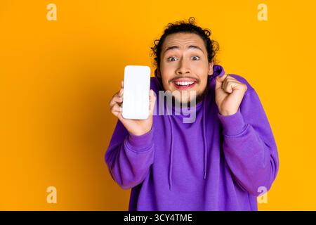 Excited young man in purple hoodie happily shows smartphone screen against yellow background Stock Photo