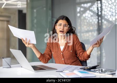 Indian businesswoman sitting at office desk feeling overwhelmed and stressed by many financial documents, holding papers in hands and having a confused expression Stock Photo