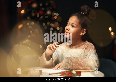 Black girl child sitting at table holding pencil to chin appearing thoughtful while drawing with colored pencils, Christmas tree with decorations glowing in background Stock Photo