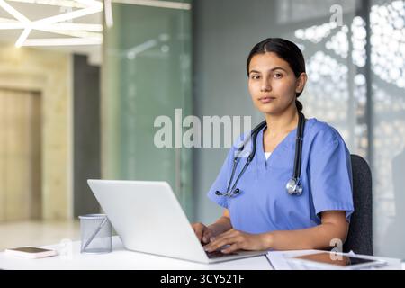 Young female healthcare professional wearing scrubs and a stethoscope, working and interacting with a laptop on a desk in a modern hospital or clinic environment Stock Photo