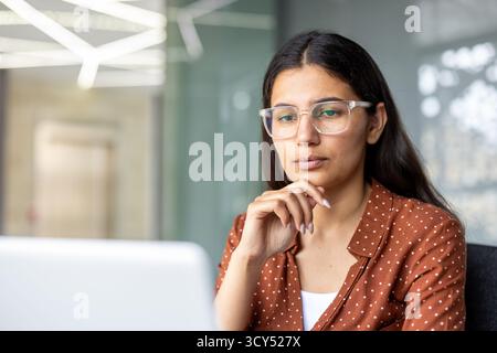 Thoughtful young woman in transparent-frame glasses concentrating on a laptop screen in a modern office, focused on work, studying or remote professional tasks Stock Photo