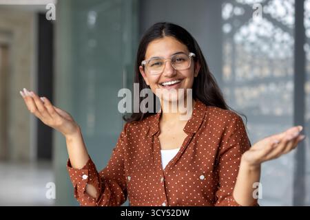 Smiling young indian woman wearing glasses and a brown polka dot shirt, standing inside and gesturing with open hands, conveying welcome, explanation, or invitation Stock Photo