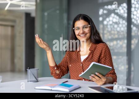 Indian businesswoman smiling at her office desk, holding a notebook and gesturing confidently while leading a meeting or presentation, mentoring colleagues and discussing strategy Stock Photo