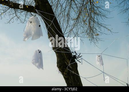 Halloween outdoor decoration featuring hanging ghost figures and a large fake spider on a web attached to a tree against a clear blue sky Stock Photo