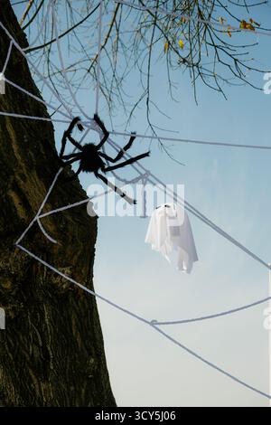 Halloween outdoor decoration with a large fake spider on a web attached to a tree and a small ghost figure hanging against a clear blue sky Stock Photo