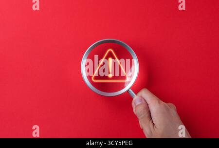 Warning alert system concept. A hand holds a magnifying glass over a glowing exclamation mark warning sign on a bold red background. hacked Stock Photo