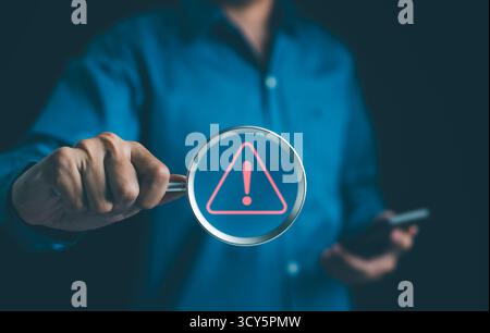 Warning alert system concept. Man holding magnifying glass focused on a glowing exclamation mark warning sign red. hack on computer network, crime Stock Photo